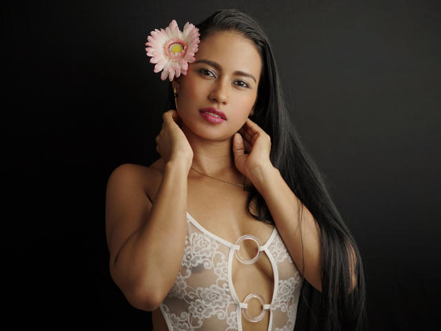 ThaliaCohen with long black hair, pink flower in hair, wearing white lace lingerie with circular cutouts, hands near neck, black background, sexy, small tits, light brown skin.