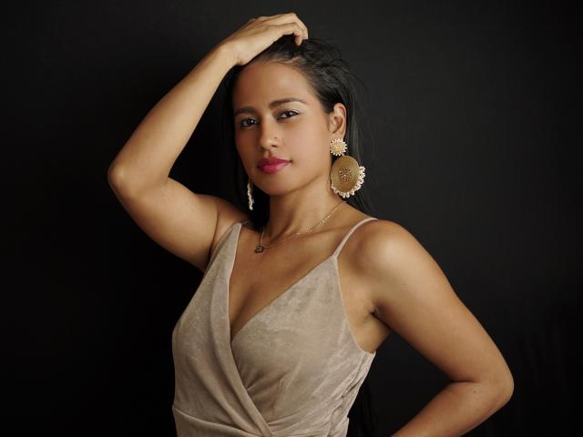 Thalia Cohen hat dunkles Haar, gebräunte Haut und rosafarbenen Lippenstift. Sie trägt ein beiges Kleid mit tiefem Ausschnitt, goldene Ohrringe und eine Halskette. Ihre Hand ist in ihrem Haar..