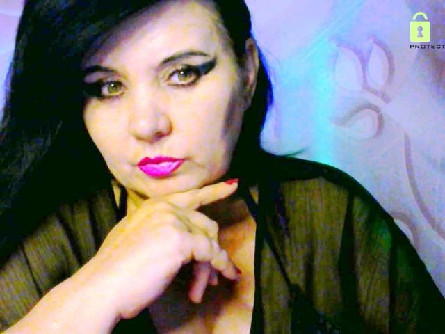 KamilaDream tem cabelo escuro, olhos verdes e batom rosa, vestindo uma blusa preta transparente com o dedo tocando o queixo. Ela parece sexy e confiante..