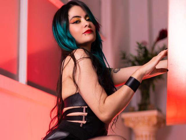 CandiceValo, avec ses cheveux turquoise, son rouge à lèvres rouge, sa robe noire sans bretelles, ses tatouages apparents sur le côté, son bracelet noir, appuyée contre un mur rouge, regardant par-dessus son épaule, éclairage rouge doux, cheveux noirs longs..