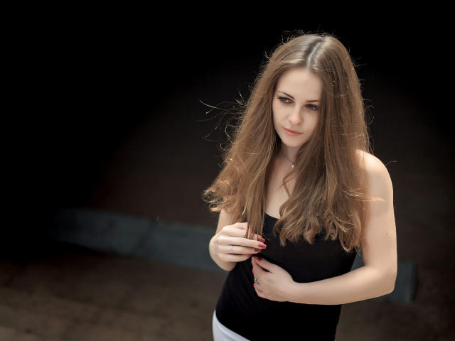 LuckyRich with long brown hair, wearing a black strap top, touching her chest with one hand, looking slightly annoyed, standing in a dark, blurred background.