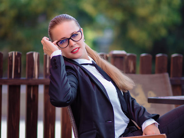 LuckyRich with slicked-back blonde hair, black glasses, red lipstick, black suit, white shirt, black tie, leaning on wooden fence, right hand in hair, confident smile.