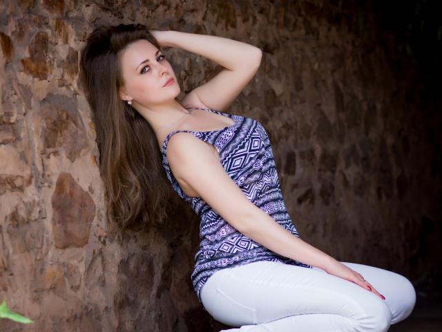 LuckyRich with long brown hair, wearing a purple and black patterned tank top, white pants, and earrings, kneeling against a stone wall, hand in hair, looking to the side.