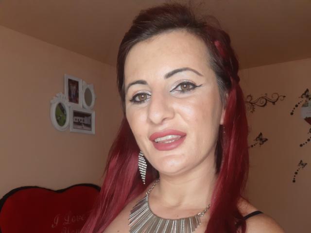 NatalyaRosse with red hair, wearing a metallic necklace, has brown eyes, winged eyeliner, red lipstick, and a slight smile, showing her tits in a selfie.