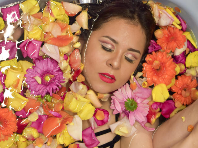 Il viso di Katie Woolf è circondato da petali di fiori colorati immersi nell'acqua, gli occhi sono chiusi con palpebre rosa, indossa un rossetto rosso e un top nero a spalline, appare rilassata e serena..