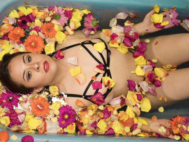 Katie Woolf, allongée dans une baignoire bleue remplie d'eau et de pétales de fleurs colorés, porte de la lingerie noire à bretelles fines, avec du rouge à lèvres et une expression calme. Ses seins sont visibles..