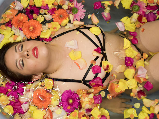 Katie Woolf, acostada en una bañera llena de agua, con pétalos de flores de colores cubriendo su bikini negro con tiras, lápiz labial rojo y cabello castaño, mirando hacia arriba con una expresión relajada y sensual..