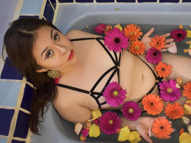 Katie Woolf en una bañera llena de agua y flores coloridas, vistiendo lencería negra con tirantes, mirando hacia arriba con lápiz labial rojo y aretes amarillos, mostrando sus senos y su abdomen suave..