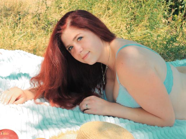 JessikaDaniels with red hair, lying on a blue-striped blanket, wearing a light blue bra, smiling at the camera, hands near her tits, sun hat and apple in the foreground.