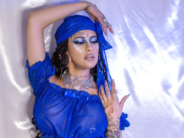 MollyWaxx, con un seno molto prosperoso, indossa un abito blu trasparente, un foulard e gioielli d'argento, ha i capelli intrecciati, ombretto blu e tatuaggi, ed è in posa con le braccia alzate davanti a uno sfondo lucido..