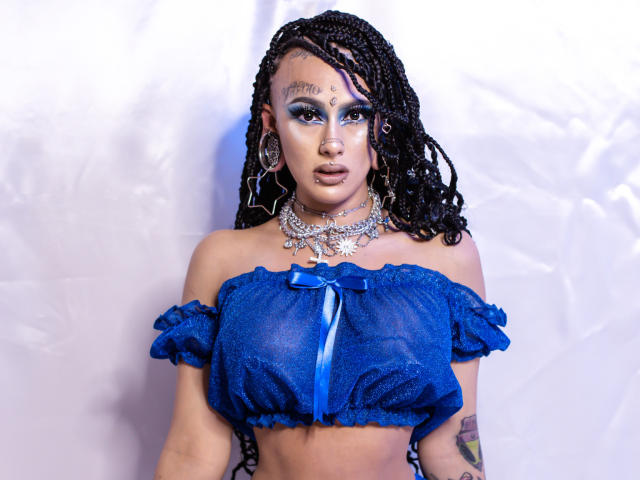 MollyWaxx con i capelli neri intrecciati, una maglia blu trasparente con una spalla scoperta, un nastro blu, grandi orecchini a stella, una collana d'argento, trucco e un anello al naso, che mostra i suoi seni su uno sfondo bianco..