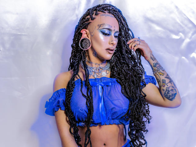 MollyWaxx a de longues tresses noires, un haut transparent bleu qui laisse entrevoir ses seins, des tatouages, du maquillage bleu pour les yeux, de grandes boucles d'oreilles et un anneau de nez, et elle se tient devant un fond blanc..