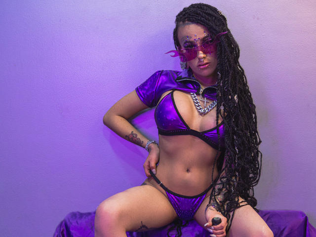 MollyWaxx, avec de longues tresses noires, un bikini violet brillant et un haut assorti, portant des lunettes roses et un collier imposant, assise les jambes écartées, montrant ses seins et ses fesses..
