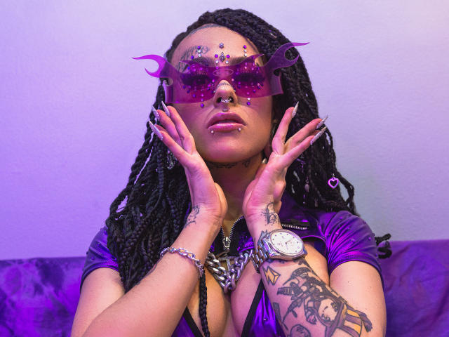 MollyWaxx con maschera trasparente viola, capelli intrecciati, anello al naso, piercing al labbro, tatuaggi e una maglietta viola, mani sul viso, orologio al polso, illuminazione viola, unghie rosa e braccialetto rosa..