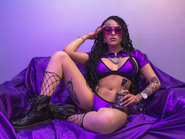 MollyWaxx con costume da bagno bikini viola metallizzato, stivali neri, calze a rete, capelli intrecciati, che tiene una bottiglia, indossa occhiali da sole rosa, con tatuaggi visibili, seduta su un tessuto di raso viola..