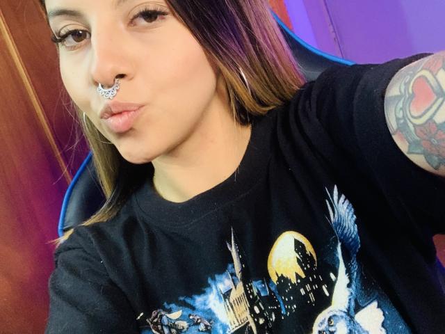 SweetAnaFlowers, com um piercing no nariz, lábios carnudos, camiseta preta e tatuagem no braço direito, usando brincos de argola, em um selfie. Ela tem um visual fofo e sexy..