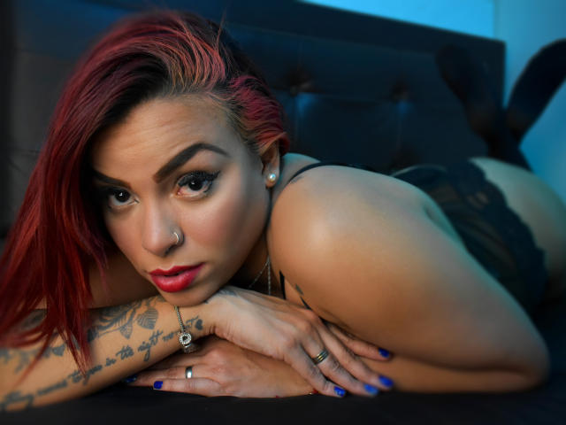 AngelFallon with red-dyed hair, lying on a black bed, wearing black lace lingerie, showing tattoos, blue nail polish, and looking at the camera with red lipstick and nose ring.