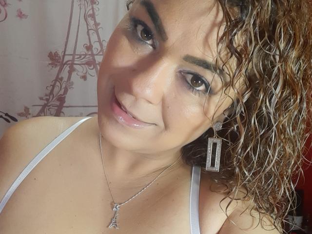 SolmaryHot has curly brown hair, medium skin, and is smiling with dark eyeliner. She's wearing a white top and silver earrings with a small pendant, showing her tits.