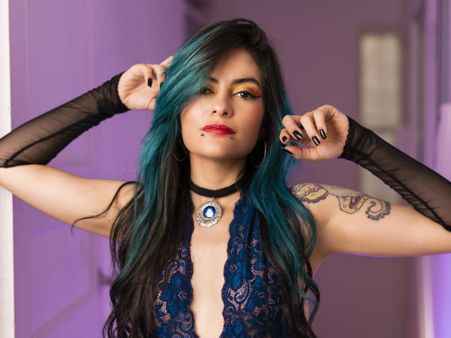 CandiceValo with teal-black hair, lace blue dress, black arm sleeves, tattoo on right arm, blue pendant, red lipstick, and black nails, hands in hair, looking at camera.