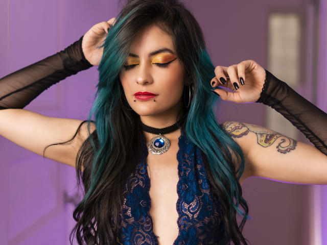 CandiceValo with teal-black hair, gold eyeshadow, red lipstick, black choker, lace top, blue gem, tattoos, black gloves, hands in hair, black nails, purple light.