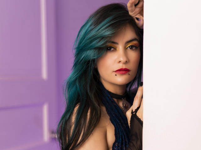Candice Valo, con cabello verde azulado y negro, lápiz labial rojo, collar negro y perforación en los pezones, asomándose desde una pared blanca, mostrando sus senos y una mirada seductora..