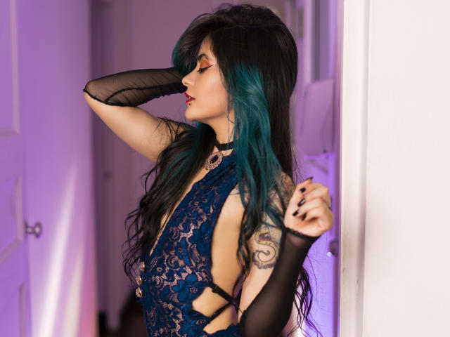 CandiceValo with long teal and black hair, wearing a lace blue dress, black arm sleeves, and red lipstick, has a tattoo on her right arm, standing in a doorway.