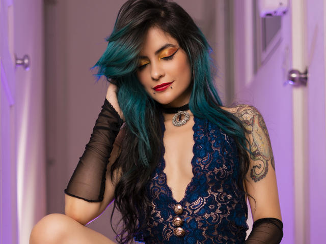 Candice Valo, com cabelo azul-turquesa, vestido azul rendado, luvas pretas, braço tatuado, batom vermelho, colar e vestido abotoado, olhos fechados, mão no cabelo, aparência sexy..