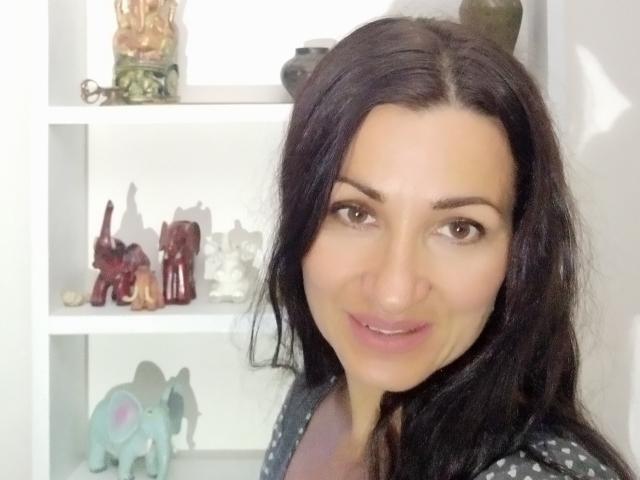 GinaONeon has dark brown hair, brown eyes, and a subtle smile. She's wearing a polka dot shirt, with small, colorful animal figurines behind her.