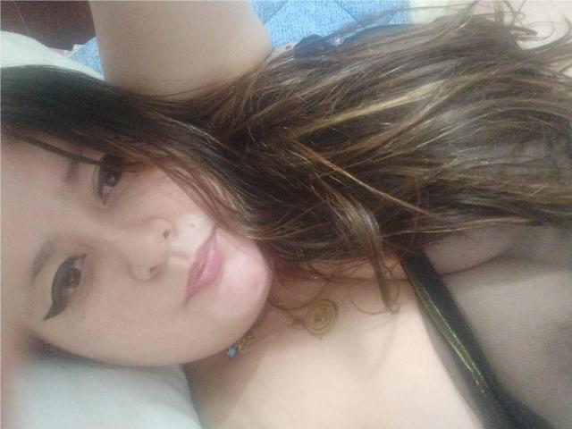 HotValeryPassione, con i capelli castano ondulati, l'eyeliner e le labbra rosa, sdraiata su un letto, indossa un reggiseno nero con spalline, mostrando i suoi piccoli seni e una collana con un ciondolo rotondo..
