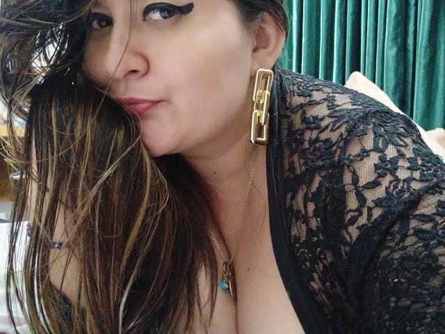 HotValeryPassione with long brown hair, black lace top, gold earrings, and thick black eyeliner, leaning in with pouty lips, showing a bit of her tits.