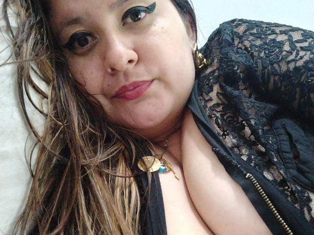HotValeryPassione with brown hair, black lace top, and gold necklace, showing her tits, wearing red lipstick, and black eyeliner, lying down, looking at the camera.