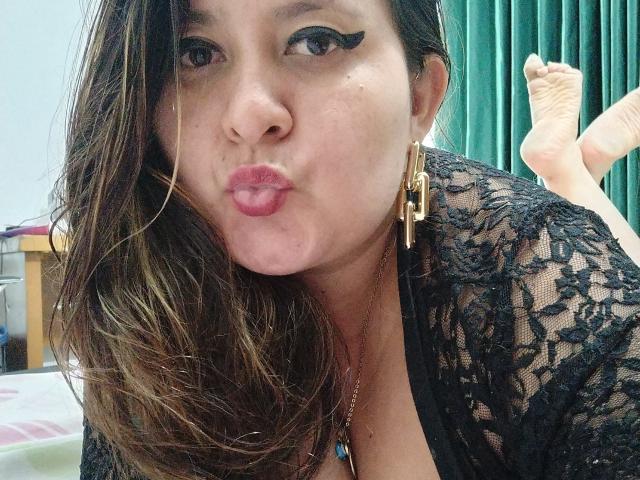 HotValeryPassione con labbra piene, eyeliner nero e orecchini dorati, indossa pizzo nero, i capelli castani le ricadono sulle spalle, le gambe sollevate, mostrando le piante dei piedi, in una posa sexy e rilassata..