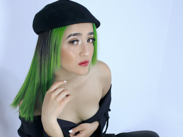 SalomePrince con i capelli verdi, un berretto nero e una maglietta nera con una spalla scoperta, che mostra il seno, guarda la fotocamera con rossetto rosso e smalto bianco..