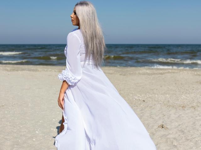 TanyaMonroe in a white, semi-transparent dress with ruffled sleeves, standing on a sandy beach, facing away, showing her ass, with long, straight, silver hair, ocean in background.