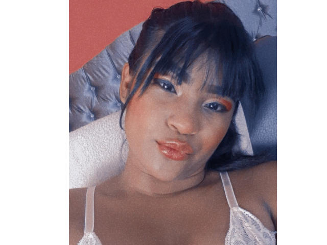Leyza hat dunkelblaues Haar, eine Pony und orangefarbene Eyeliner. Sie liegt auf einem blauen, gepolsterten Kopfstütze, verzogen den Mund mit glänzlichen Lippen und trägt eine weiße Spitzenbluse..