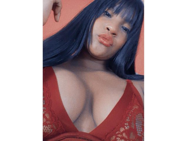 Leyza, com cabelo azul, lábios laranja e um sutiã de renda vermelha que mostra seus seios grandes e arredondados. Ela está olhando para baixo, com uma mão perto da cabeça..