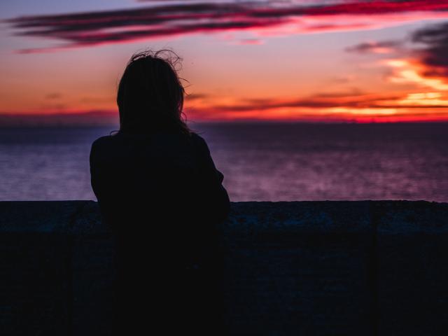 Silhouetted FairyRoni with messy hair stands against a stone wall, back to the camera, watching a vivid sunset over a dark purple ocean, hands by their side.