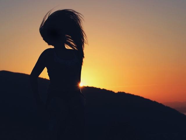 Eine Silhouette von FairyRoni mit fließendem Haar, mit dem Rücken zur Kamera, Sonnenuntergang im Hintergrund, trägt ein Tanktop, das Gesäß ist leicht angehoben, die Sonne lugt am Horizont hervor, der Himmel ist orange und gelb..