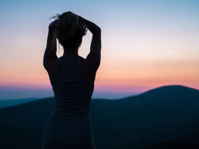 Silhouet van FairyRoni met armen omhoog, gekleed in een gestreepte jurk, staand voor een zonsondergang. Haar achterkant is zichtbaar, en haar haar zit in een rommelige knot..