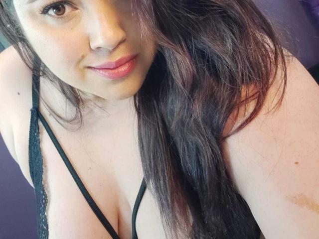 LissaClaire with long dark brown hair, pink lips, and black strappy lingerie, showing her large tits and a slight smile. Her brown eyes and fair skin are visible.