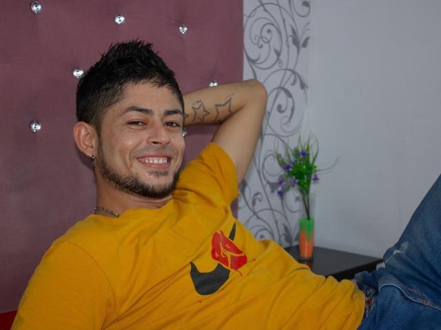 AnthonyHotyBig, klein, donker haar, lichte baard, geel shirt met een rood embleem, tatoeage op de rechterarm, ontspannen, lachend, blauwe jeans, arm achter het hoofd, zittend, casual, sexy..