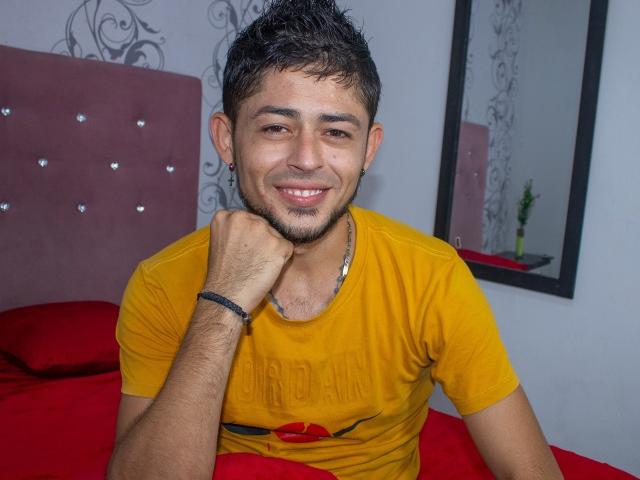 Anthony Hoty Big, con cabello oscuro y corto, una ligera barba, sonriendo, camiseta amarilla, cama roja, con el mentón apoyado en la mano, pulsera en la muñeca, aretes negros, espejo en la pared, almohadas rojas..