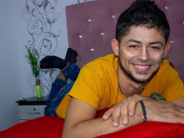 AnthonyHotyBig, deitado em uma cama vermelha, sorrindo, com cabelo curto e escuro, pele clara, camisa amarela e pulseira preta. Os pés aparecem ao fundo, levemente visíveis..