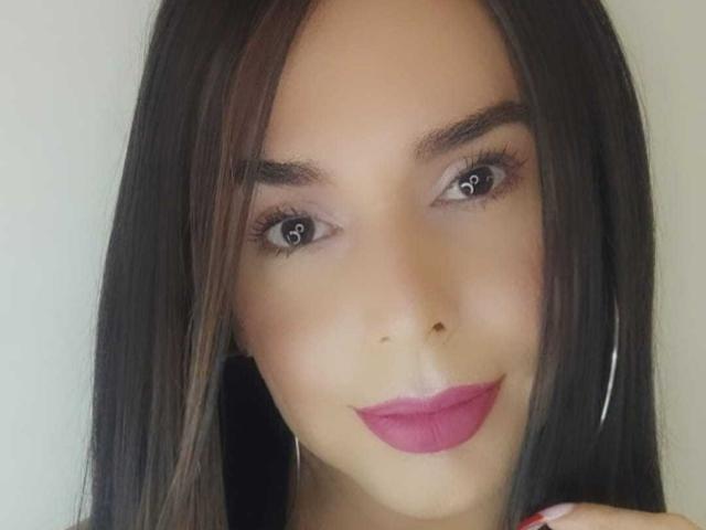Jhovanna has dark brown eyes with heart-shaped pupils, straight black hair, pink lipstick, and wears silver hoop earrings. Her skin is tan and she's smiling softly.