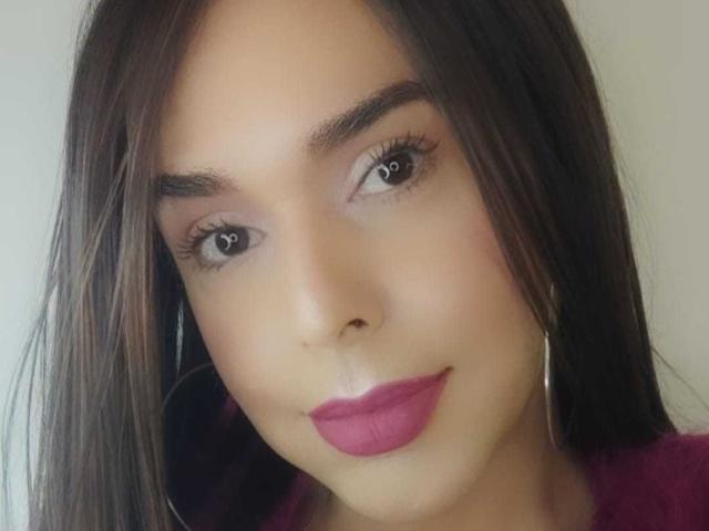 O rosto de Jhovanna, com cabelo castanho escuro, batom rosa, cílios longos e pequenas argolas nos ouvidos, mostrando um sorriso discreto. Sua pele é bronzeada e ela tem sobrancelhas bem cuidadas..