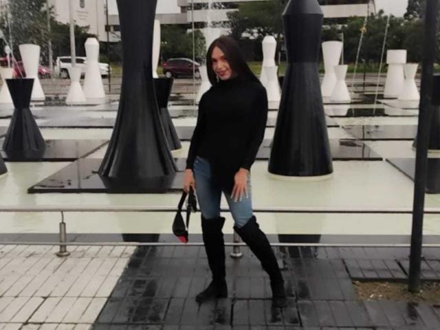 Jhovanna mit mittelgroßer Brust, einem schwarzen Pullover, blauen Jeans und schwarzen Kniehochstiefeln, hält eine schwarze Tasche und steht vor hohen, schwarz-weißen Skulpturen..