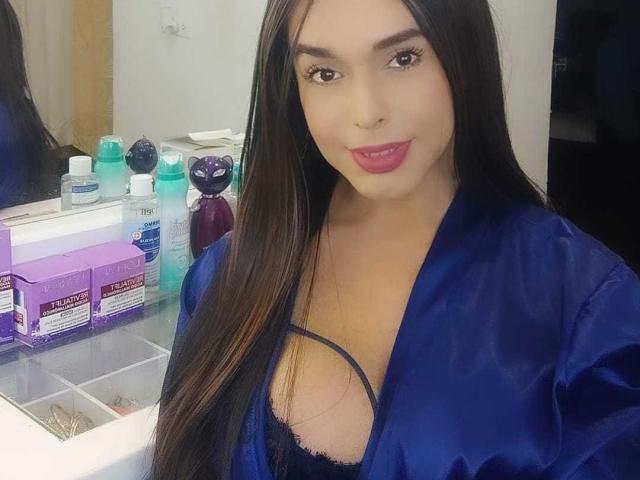 Jhovanna with long black hair, wearing a blue satin robe, exposing her large tits in a black lace bra, smiling with pink lipstick, standing in front of a makeup counter.