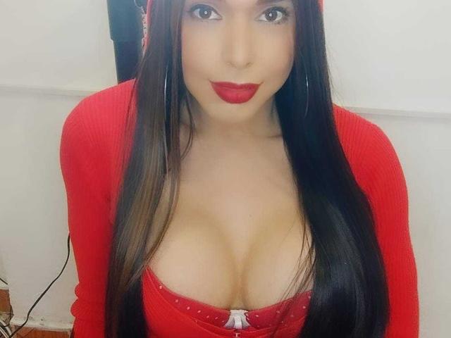 Jhovanna with long black hair, red cardigan, and red lipstick, showing her big tits in a red top with a white bow. She's looking at the camera with a smile.