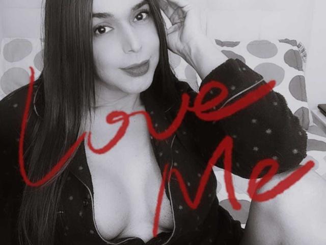 Jhovanna, avec ses longs cheveux noirs, portant une robe noire à pois, légèrement ouverte pour dévoiler ses seins, avec l'inscription cursive rouge "aime-moi" sur sa poitrine, assise sur un coussin à motifs..