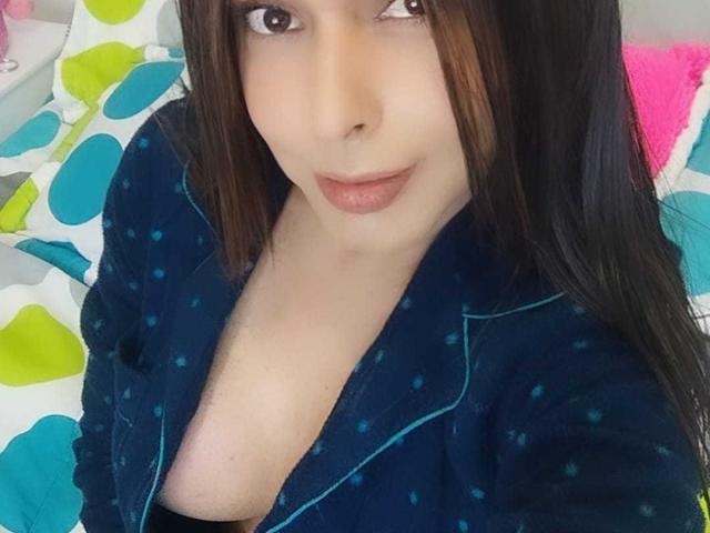 Jhovanna, con ojos marrones, cabello largo y oscuro, y labios rosados, vestida con una bata azul de lunares, mostrando parte de su escote, mirando hacia la cámara. Sus senos son parcialmente visibles..